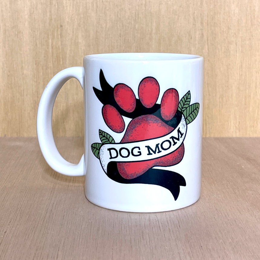 white coffee mug photographed against a wood grain background. The mug has a tattoo -style design featuring dog's paw print in red, with a banner flag wrapping around it that reads 'dog mom'. There are green leaves accenting the design.