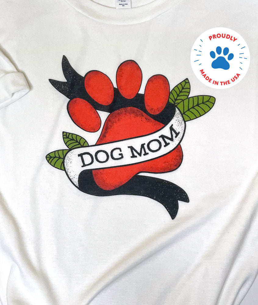 white t-shirt photographed flat, it has a design of Dog Mom with paw print graphic in a tattoo style, proudly made in the USA