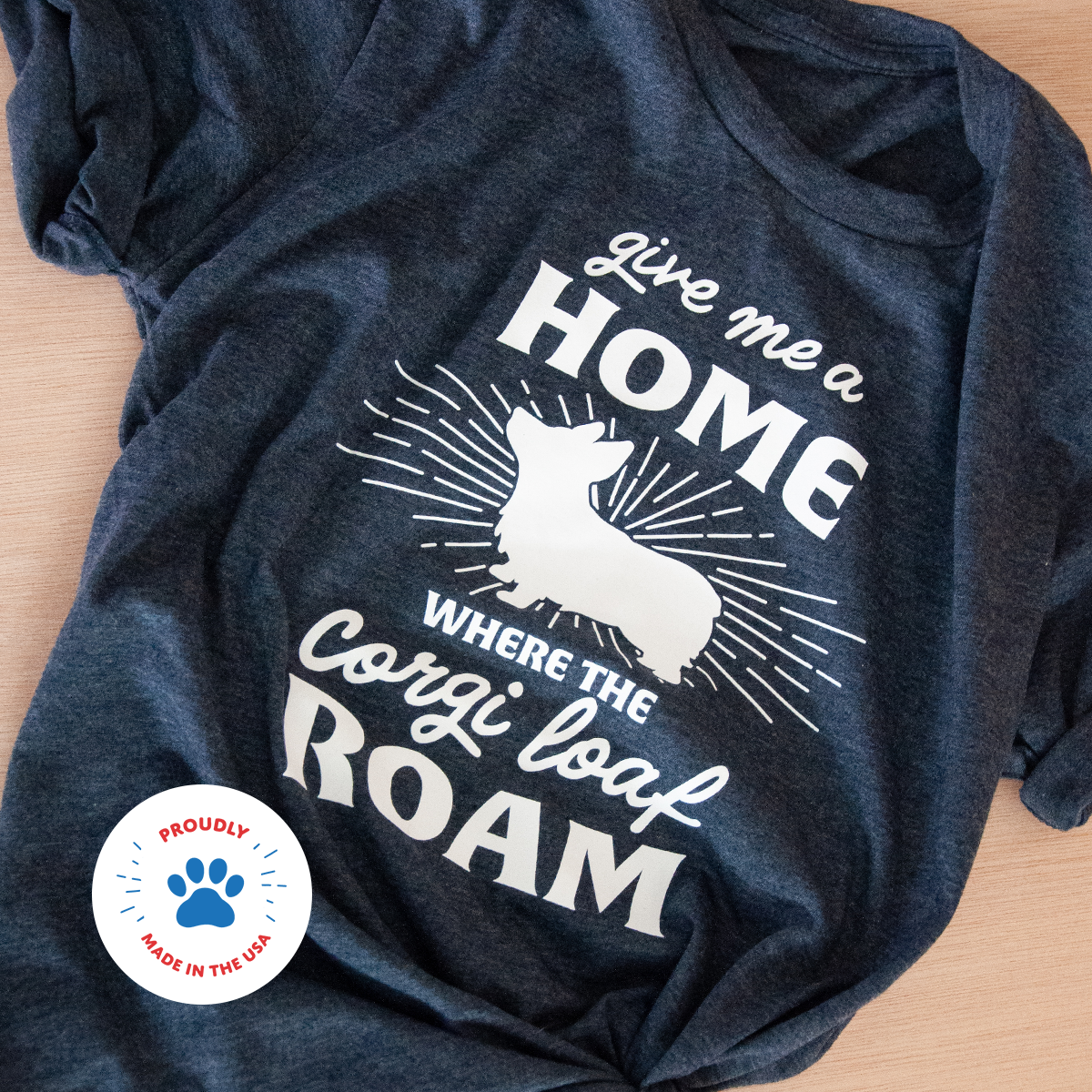 T-shirt lays flat on wood. Shirt is navy blue. Design is white. Design reads Give Me a Home Where the Corgi Loaf Roam.  Made in the USA.