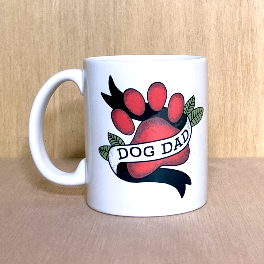 white coffee mug photographed against a wood grain background. The mug has a tattoo -style design featuring dog's paw print in red, with a banner flag wrapping around it that reads 'dog dog'. There are green leaves accenting the design.