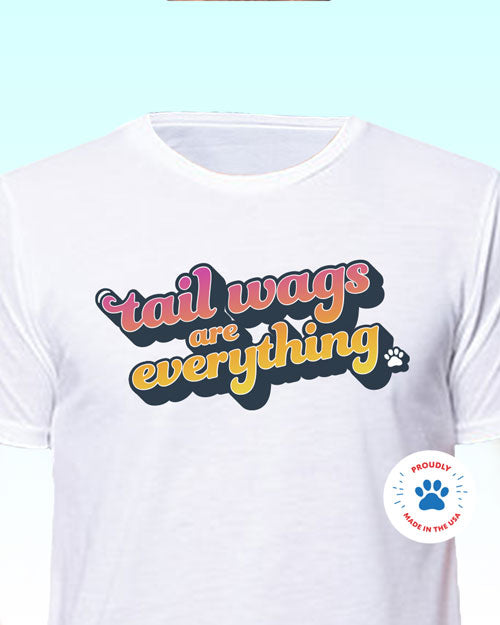 A white t-shirt with a bright pink and yellow retro style text design, which reads: Tail Wags Are Everything. Paw print graphic on shirt. Proudly made in the USA paw print icon overlay.